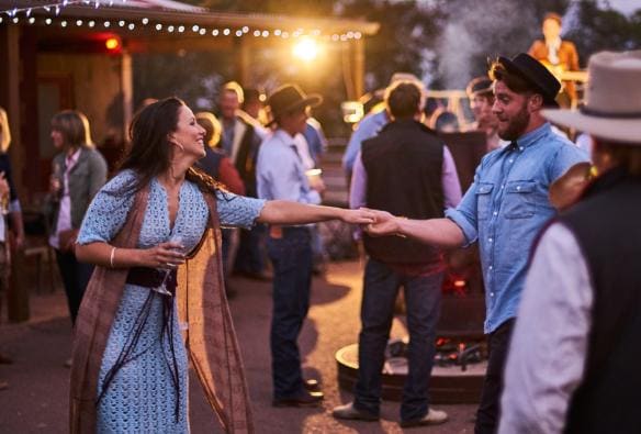 Un uomo e una donna che si tengono per mano mentre ballano tra altre persone all'esterno di un locale al tramonto al Prairie Hotel, Flinders Ranges & Outback, South Australia © South Australian Tourism Commission