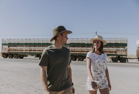 Un uomo e una donna che camminano su una pianura sabbiosa dell'outback con un grande camion dietro di loro al Blue Heeler Hotel, Kynuna, Queensland © Tourism and Events Queensland