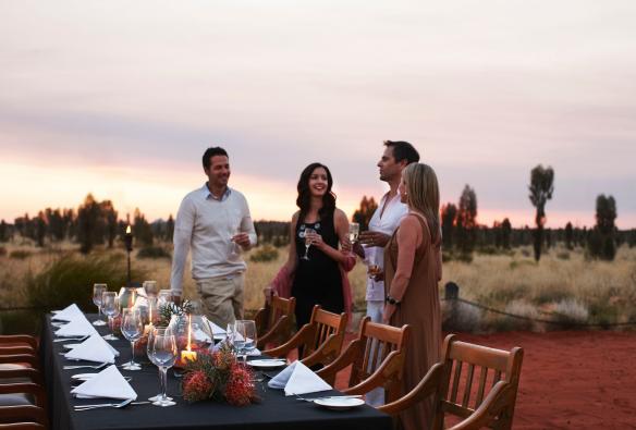 Quattro persone con bicchieri di vino intorno a un tavolo all'aperto decorato con candele e fiori autoctoni nel deserto presso Table 131, Longitude 131, Uluru Kata Tjuta National Park, Northern Territory © Luxury Lodges of Australia