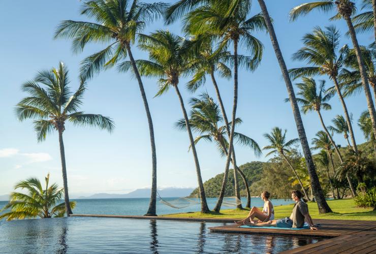 Coppia seduta sugli asciugamani accanto alla piscina a sfioro con vista sull'oceano con palme sullo sfondo presso l'Orpheus Island Lodge, Orpheus Island, Queensland © Tourism and Events Queensland