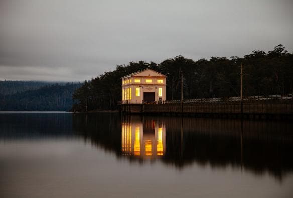 Accomodation di Pumphouse Point illuminata al calar della notte, riflessa insieme al panorama nelle acque tranquille di Lake St Clair, Tasmania © Tourism Tasmania