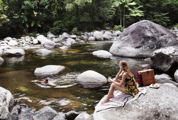 Donna seduta su un masso liscio con un bicchiere di vino e un cesto da picnic, che osserva un uomo nuotare nelle acque limpide del Mossman Gorge River vicino al Silky Oaks Lodge, Queensland © Luxury Lodges of Australia