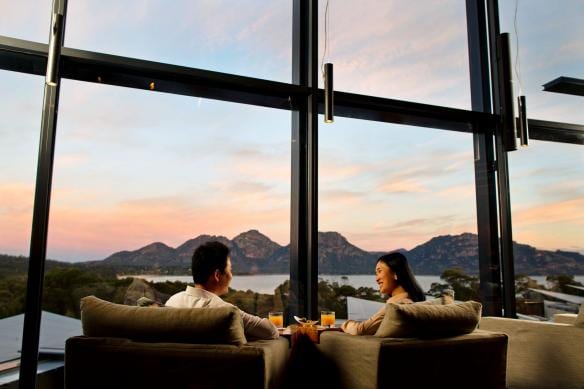 Una coppia cena accanto a grandi finestre a tutta altezza che si affacciano su una baia calma circondata da montagne rocciose, Saffire Freycinet, Freycinet, Tasmania © Tourism Australia