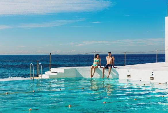 Un uomo e una donna seduti sul bordo della piscina di Bondi Icebergs con i piedi nell'acqua e l'oceano alle loro spalle a Sydney, New South Wales © Destination NSW