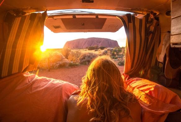 Una persona sdraiata nel retro di un furgone con il portellone aperto per ammirare la vista su Uluru durante un'alba dalle tonalità dorate, Uluru-Kata Tjuta National Park, Northern Territory © Mitchell Cox /Tourism NT