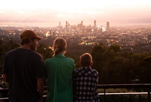 Una famiglia in piedi al Mount Coot-Tha Lookout che ammira le viste sullo skyline della città e sugli edifici illuminati a Brisbane, Queensland © Tourism and Events Queensland