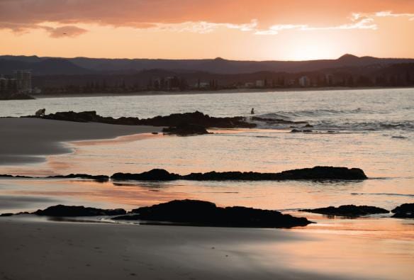 Acque poco profonde che riflettono il cielo rosa all'alba su una distesa di sabbia piatta a Snapper Rocks con un surfista che cavalca le onde nell'oceano, Gold Coast, Queensland © Tourism and Events Queensland