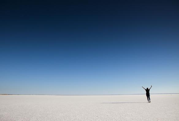 Persona in piedi sul fondo di una salina rosa chiaro con le braccia alzate in aria a Lake Hart, outback del South Australia © FROSAT & Singing Bowl Media