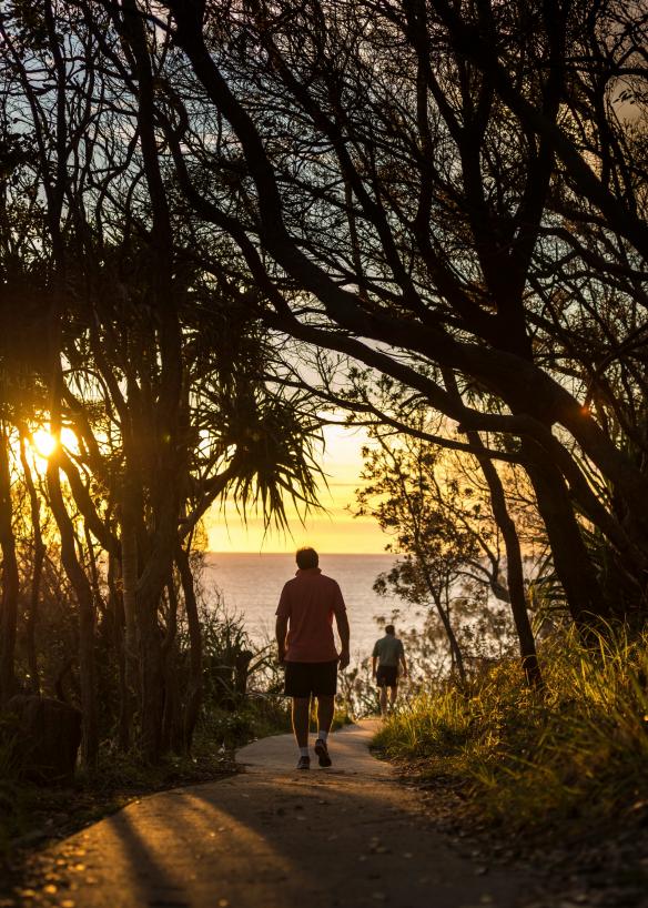 Persone che camminano lungo un sentiero nel bushland mentre il sole tramonta sull'oceano davanti a loro a Dolphin Point, Noosa National Park, Noosa, Queensland © Tourism Australia