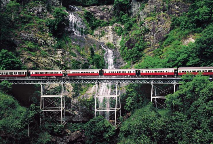 Treno che viaggia su binari sopraelevati accanto a una grande cascata nella foresta pluviale, Kuranda Scenic Railway, Kuranda, Queensland © Tourism and Events Queensland
