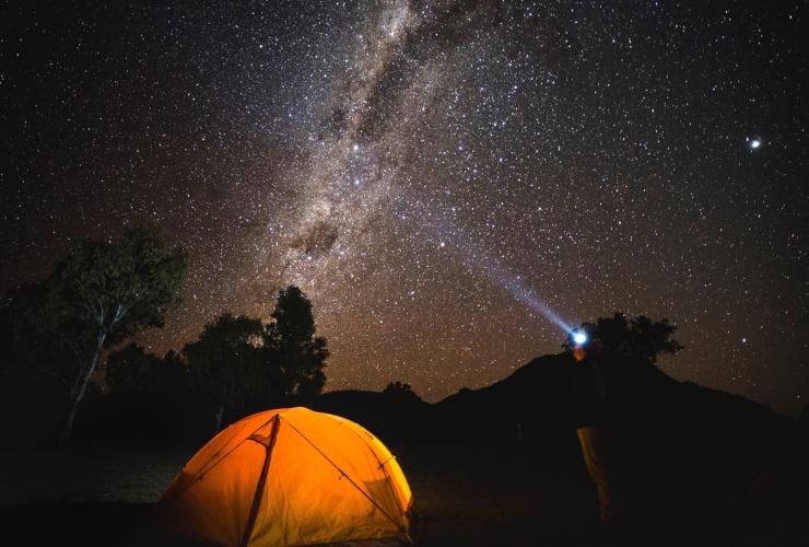 Una persona in piedi accanto a una tenda con una lampada frontale puntata verso le stelle, Dark Sky Park, Warrumbungle National Park, New South Wales © Destination NSW