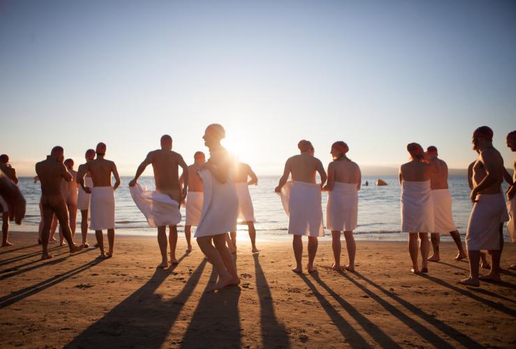 タスマニア州、ホバート、ダーク・モフォ、ヌード・ソルスティス・スイム。早朝、赤いスイムキャップをかぶりタオルを巻いた姿でビーチに立つ人々 © Dark Mofo