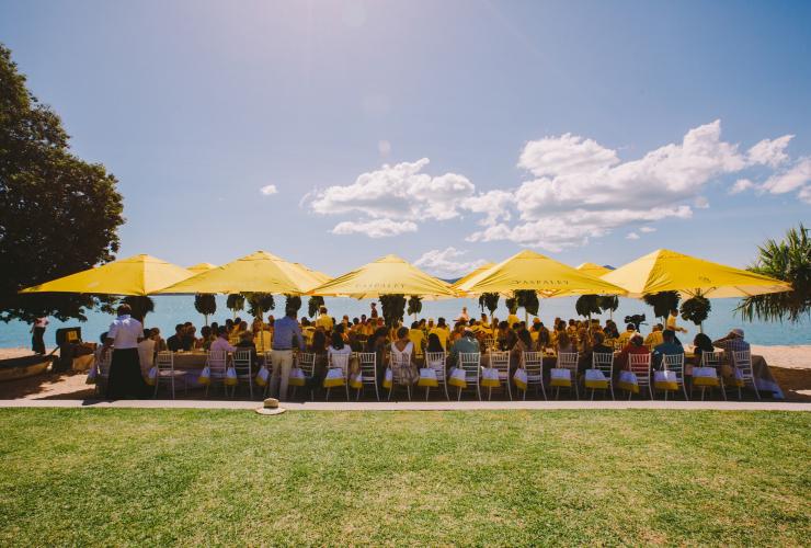 クイーンズランド州、ハミルトン島、パスパレイの「ストランデッド・アット・クオリア」ランチ。草原と青い海の間に設置した黄色いパラソルの下、長いテーブルで食事をする人々 © Tourism Australia