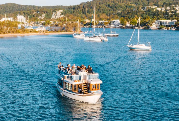 クイーンズランド州、ヌーサ、ヌーサ・イート＆ドリンク・フェスティバル。乗客を満載したボートが緑豊かな海岸から出航する様子の空撮 © Tourism and Events Queensland