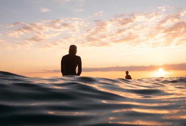 ニュー・サウス・ウェールズ州、バイロン・ベイで朝日に浮かび上がる海上でサーフボードに座っている2人のサーファーのシルエット © Tourism Australia