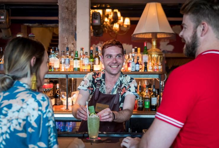 ニュー・サウス・ウェールズ州、シドニー、インペリアル・アースキンビルで、背の高いグラスの緑のカクテルを2人の客に手渡す笑顔のバーテンダー © Destination NSW