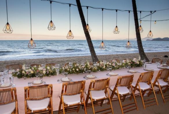 クイーンズランド州、パーム・コーブ、ヌ・ヌ・レストラン、花と吊り下げられた照明で飾られたビーチの長いテーブル © Palm Cove Weddings