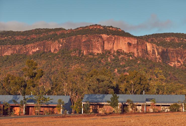 クイーンズランド州、木々に囲まれたマウント・マリガン・ロッジの宿泊施設外観と背景のマウント・マリガンの赤く険しい山々 © Mount Mulligan Lodge, Queensland