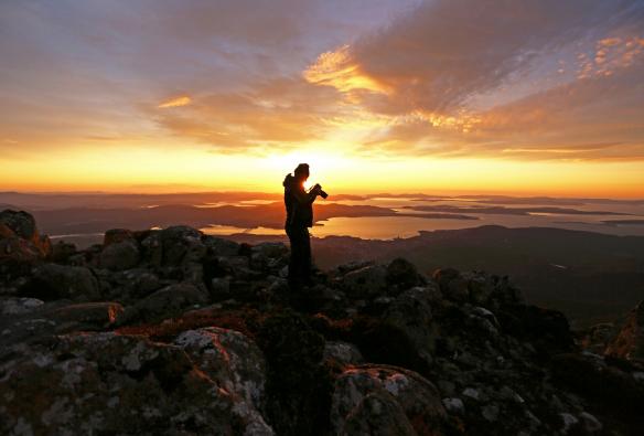 ホバートやタスマニア州周辺域に日の光が差し込む頃、クナニ/ウェリントン山の頂上で岩の上に立ち、カメラを覗き込んでいる人 © Tourism Australia