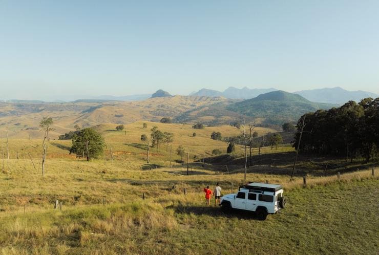 クイーンズランド州、シーニック・リム、バヤ・タイニーハウス、緑豊かなカントリーサイドで四輪駆動車の横に立つ2人 © Tourism and Events Queensland