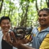 クイーンズランド州、デインツリー・レインフォレスト、ウォークアバウト・カルチュラル・アドベンチャーズ、大きなマッドクラブを掲げる先住民のガイドと、その後ろで満面の笑顔を見せるツアー参加者 © Tourism and Events Queensland