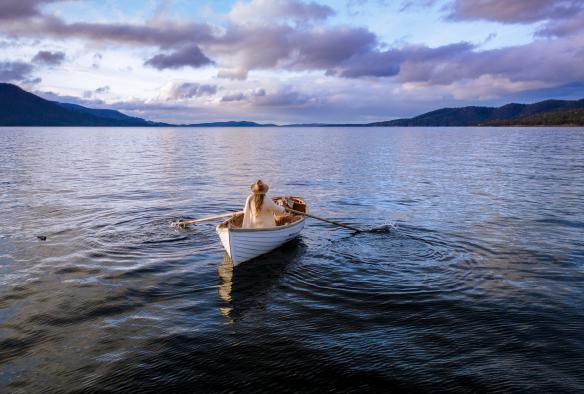 뒤로 산들이 보이는 가운데 새틀라이트 아일랜드 근처의 잔잔한 푸른 물 위에서 한 여성이 작은 나무배를 저어 가고 있는 모습, 태즈메이니아 © 애덤 깁슨