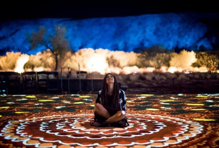 Một người ngồi trên mặt đất, xung quanh là những hoa văn ánh sáng rực rỡ đầy màu sắc tại Lễ hội Parrtjima, Thị trấn Alice Springs, Lãnh thổ phía Bắc © Tourism Northern Territory/James Horan Photography 