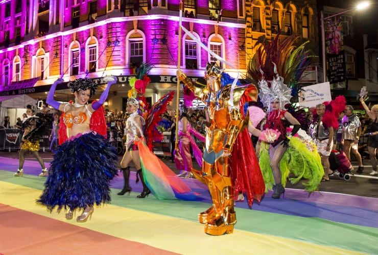 Một nhóm người đang sải bước trên con đường phủ sắc cầu vồng, khoác lên mình những chiếc váy lấp lánh ánh bạc và đội tóc giả màu vàng tại sự kiện Sydney Gay and Lesbian Mardi Gras, New South Wales © Destination NSW