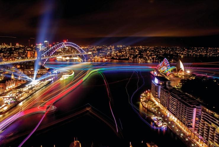 Bến cảng với một cây cầu vòm và công trình hình cánh buồm, tất cả đều được chiếu sáng bằng đèn nhiều màu trong sự kiện Vivid Sydney, Sydney, New South Wales © Destination NSW 