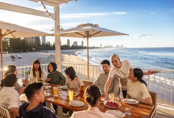 Một nhóm người ngồi ngoài trời tại bàn ăn hướng ra bãi biển, trong khi người phục vụ đặt món ăn lên bàn tại nhà hàng The Tropic, Burleigh Pavilion, Thành phố Gold Coast, Tiểu bang Queensland © Tourism Australia