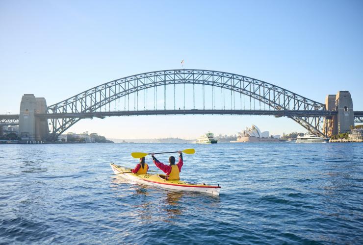 Một đôi chèo thuyền kayak giơ mái chèo lên trước Cầu Cảng Sydney tại thành phố Sydney, tiểu bang New South Wales © Tourism Australia”