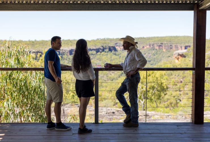 Tour guide smiling and talking with a couple holding drinks on a verandah overlooking bushland with Jarramali Rock Art Tours, Cape York, Queensland © Tourism Australia