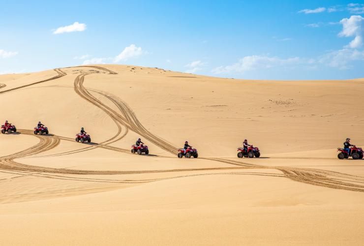 A line of people riding quad bikes across sand dunes with Sand Dune Adventures at Stockton Beach, Port Stephens, New South Wales © Destination NSW