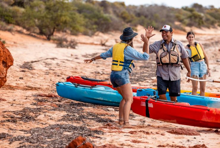An Aboriginal tour guide high fiving a guest while standing on a beach beside kayaks with Wula Gura Nyinda Eco Cultural Adventures, Coral Coast, Western Australia © Tourism Australia 
