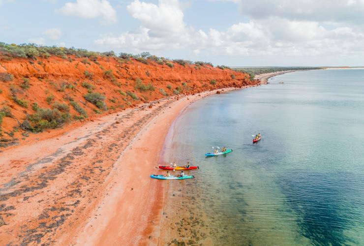 Aerial view over a group of people kayaking in clear blue water beside bright orange cliffs during a tour with Wula Gura Nyinda Eco Cultural Adventures, Coral Coast, Western Australia © Tourism Western Australia