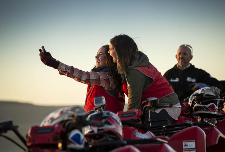 Two people taking a selfie on quad bikes during sunset with Sand Dune Adventures at Stockton Beach, Port Stephens, New South Wales © Destination NSW