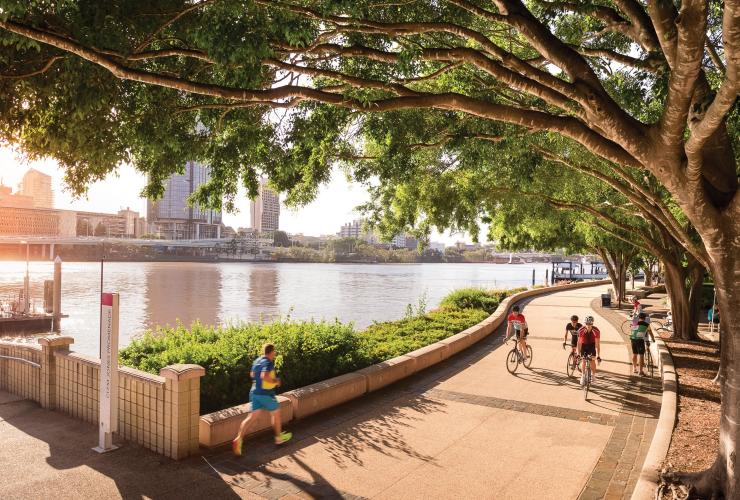 A group of people riding bikes along a leafy path beside a river with tall buildings in the background along the River Walk, South Bank, Brisbane, Queensland © Fullframe Photographics