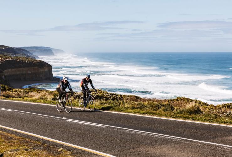Two people cycling side by side along a road beside sheer sandstone cliffs dropping down to the ocean below along the Great Ocean Road, Victoria © Belinda Van Zanen/Great Ocean Road Tourism