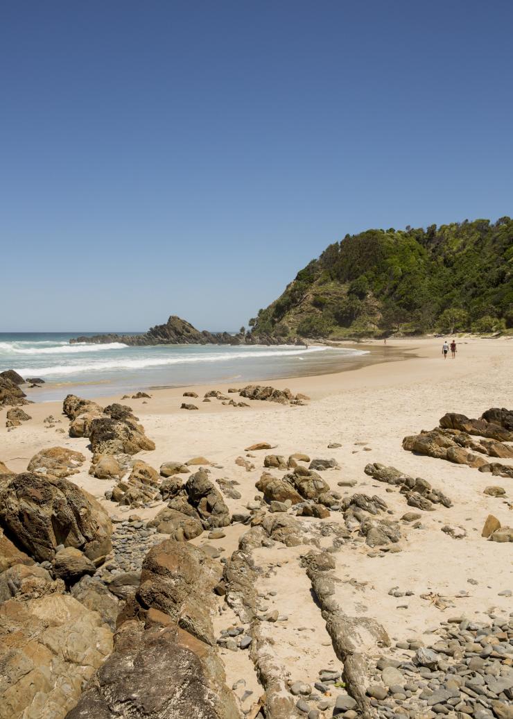 People walking on a beach with rocks leading to white sand and a green headland at Kings Beach, Byron Bay, New South Wales © Destination NSW