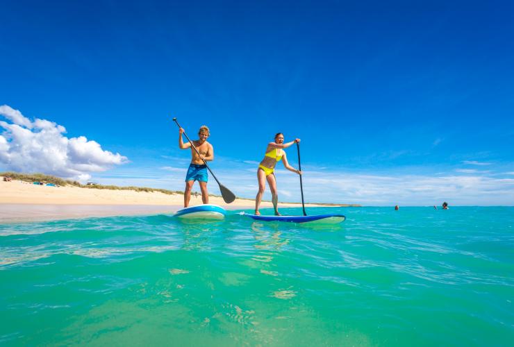 Two people stand up paddleboarding in bright blue water as two others swim in the distance at Mauritius Beach, near Exmouth, Western Australia © Tourism Western Australia