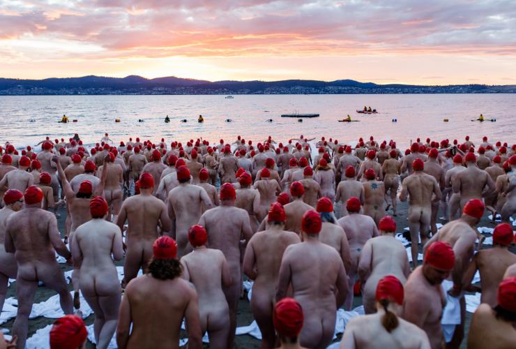 Aerial view of a crowd of nude people wearing red swimming caps running towards the calm ocean during the Nude Solstice Swim, Dark Mofo, Hobart, Tasmania © Dark Mofo/Jesse Hunniford