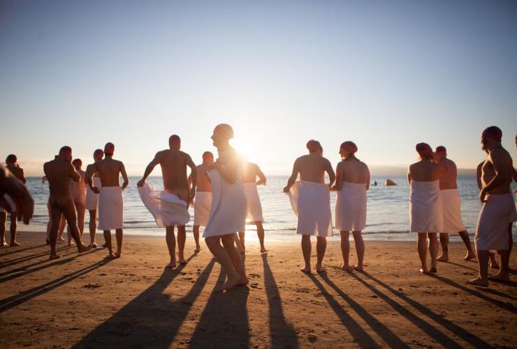 A group of people wearing white towels and red swimming caps at the edge of the ocean during the Nude Solstice Swim, Dark Mofo, Hobart, Tasmania © Dark Mofo