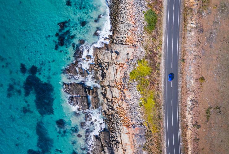 Aerial view over a car driving along a road beside orange rocks leading to clear blue ocean on the Great Eastern Drive, Freycinet, Tasmania © Tourism Australia 