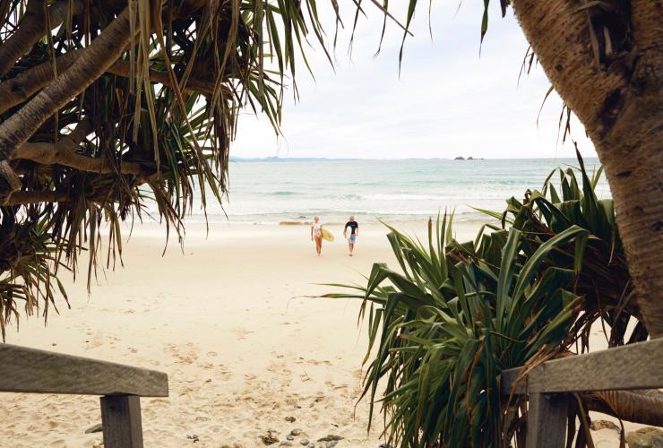 Two surfers carry surfboards under their arms as they walk across a white-sand beach toward the ocean in Byron Bay, New South Wales © Tourism Australia
