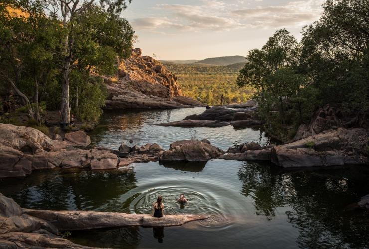 Two people swimming in a series of waterholes at the top of a cliff amid lush green bushland at Gunlom Falls, Kakadu, Northern Territory © Tourism Australia