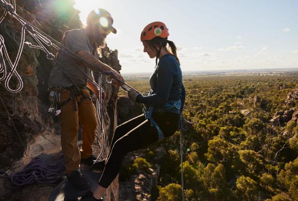 A person abseiling down a sheer cliff overlooking bushland with the help of an instructor with Hangin' Out, Grampians, Victoria © Tourism Australia 