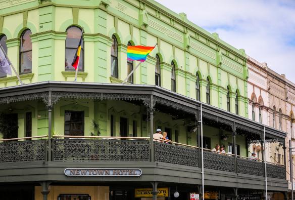 People sitting on a balcony of a large green pub with Aboriginal and LGBTQI+ flags seen blowing in the wind on the roof Newtown Hotel, Newtown, Sydney, New South Wales © City of Sydney / Katherine Griffiths