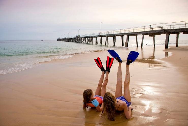 Một người mẹ và con gái đang nằm trên bãi cát, chân mang chân vịt giơ lên không tại Bãi biển Port Noarlunga, Tiểu bang Nam Úc © South Australian Tourism Commission/Adam Bruzzone