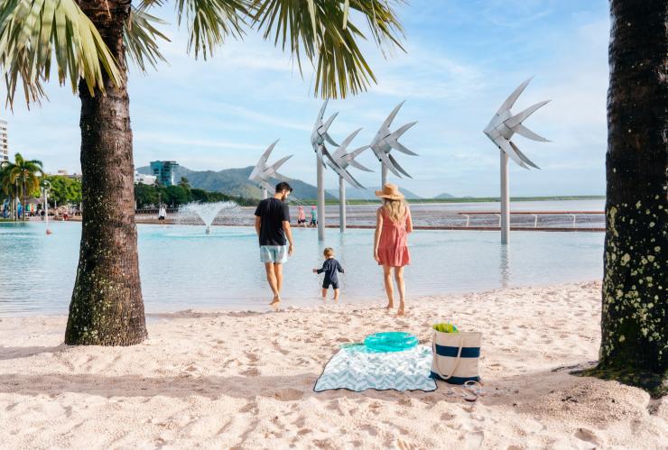 Cha mẹ và một em bé đang nắm tay nhau trên bãi biển cát trắng bên cạnh một đầm nước kín gió tại Đầm phá Cairns Esplanade, Thành phố Cairns, Tiểu bang Queensland © Tourism and Events Queensland