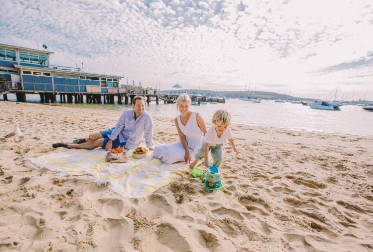 Một cặp cha mẹ và một đứa trẻ đang ngồi trên bãi biển, gần đó có thuyền neo đậu tại Bãi biển Balmoral, Thành phố Sydney, Tiểu bang New South Wales © Tourism Australia